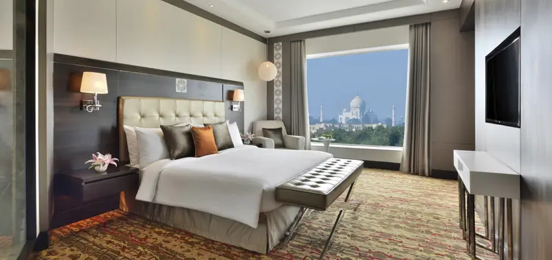 Luxurious hotel bedroom featuring a king-sized bed and a large window with a view of the Taj Mahal